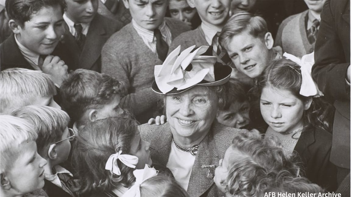 Helen as an older adult in a small hat is smiling while surrounded by a big group of children