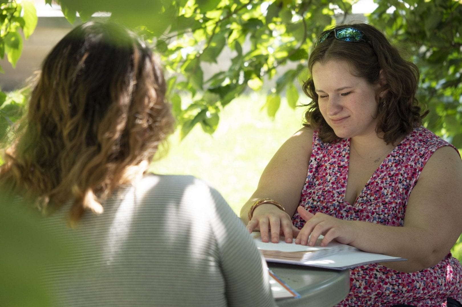 Two women sitting at a table under a tree. One is reading braille.