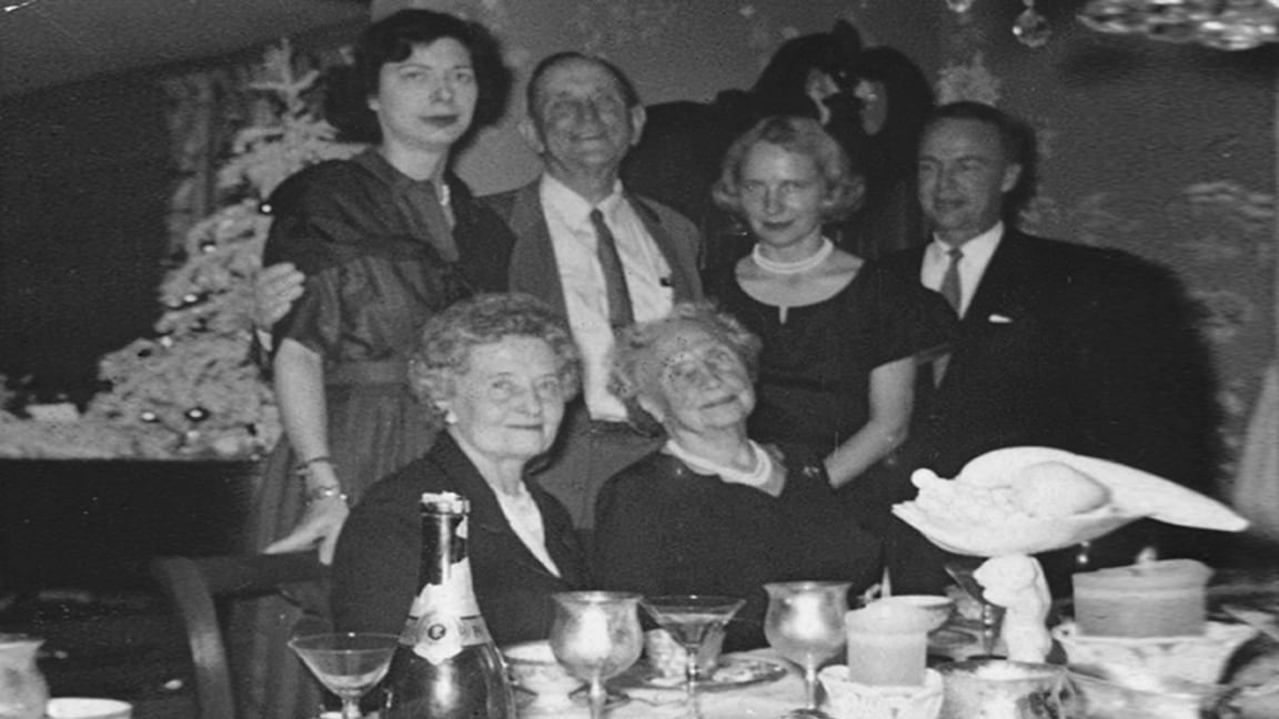 Helen is sitting at a dining table. She is wearing pearls and a dark-colored dress. Polly Thomson sits to her right; four others stand behind her, with arms around each other. A white Christmas tree decorated with ball-shaped ornaments is in the background. The table is crowded with napkins, candles, coffee cups, long-stemmed glasses, and a bottle of champagne.