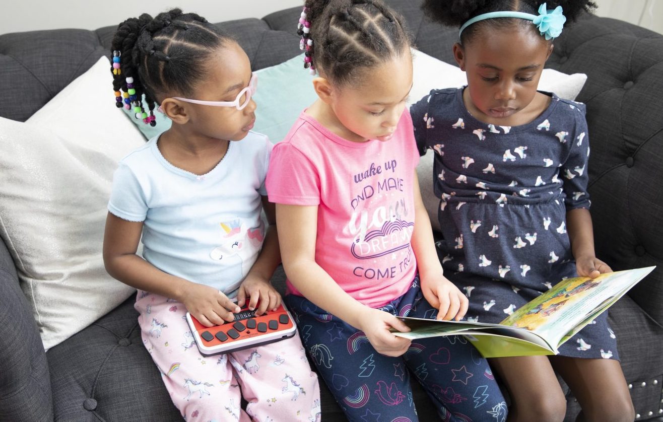 three girls reading on a couch. two are looking at a print book and one is using the braille trail reader