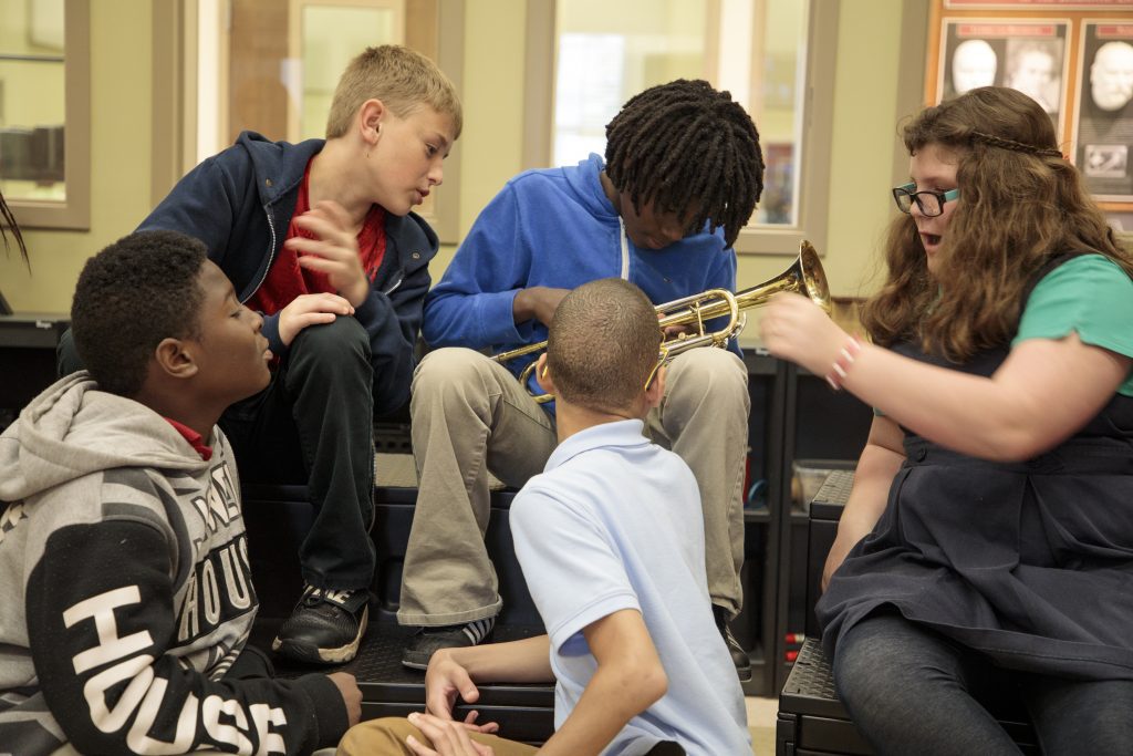 a group of students sitting together in a music classroom talking. one of them holds a trumpet.