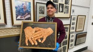 A man wearing a sweater and a baseball cap holds the same painting. He is wearing blue latex gloves to protect the painting from fingerprints. On the wall behind him hang various framed photos of Helen Keller. 