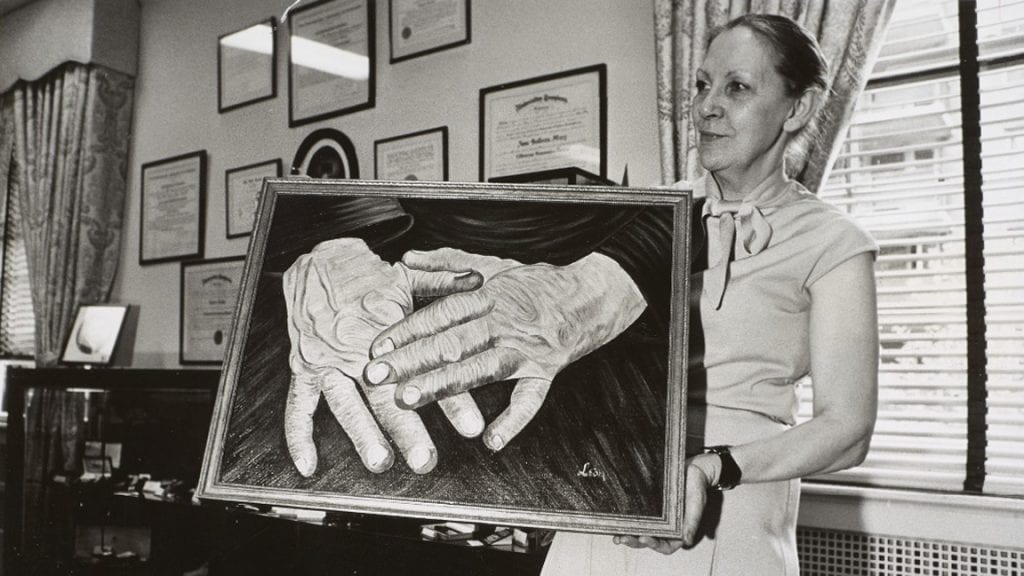 A woman holds a large painting depicting two hands, the left hand resting on the right. The woman wears a blouse with a bow and her hair has been pulled back into a bun. On the wall behind her are various framed documents.