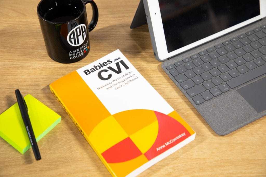 Babies with CVI book sits atop a desk, next to a laptop, APH mug, and pad of paper and pen