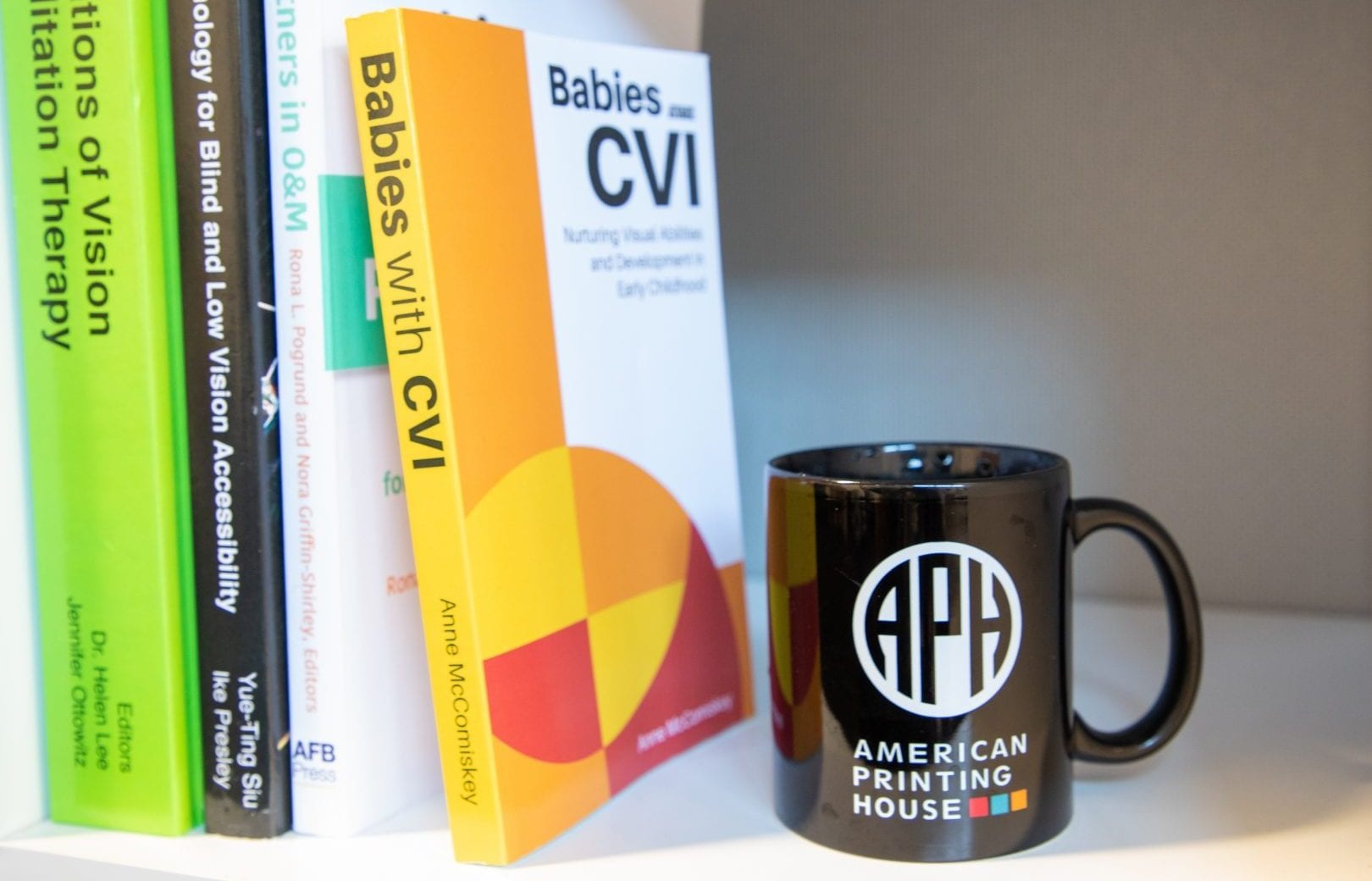 babies with CVI book sits on a bookshelf with other APH Press books. a black APH coffee up sits next to them.
