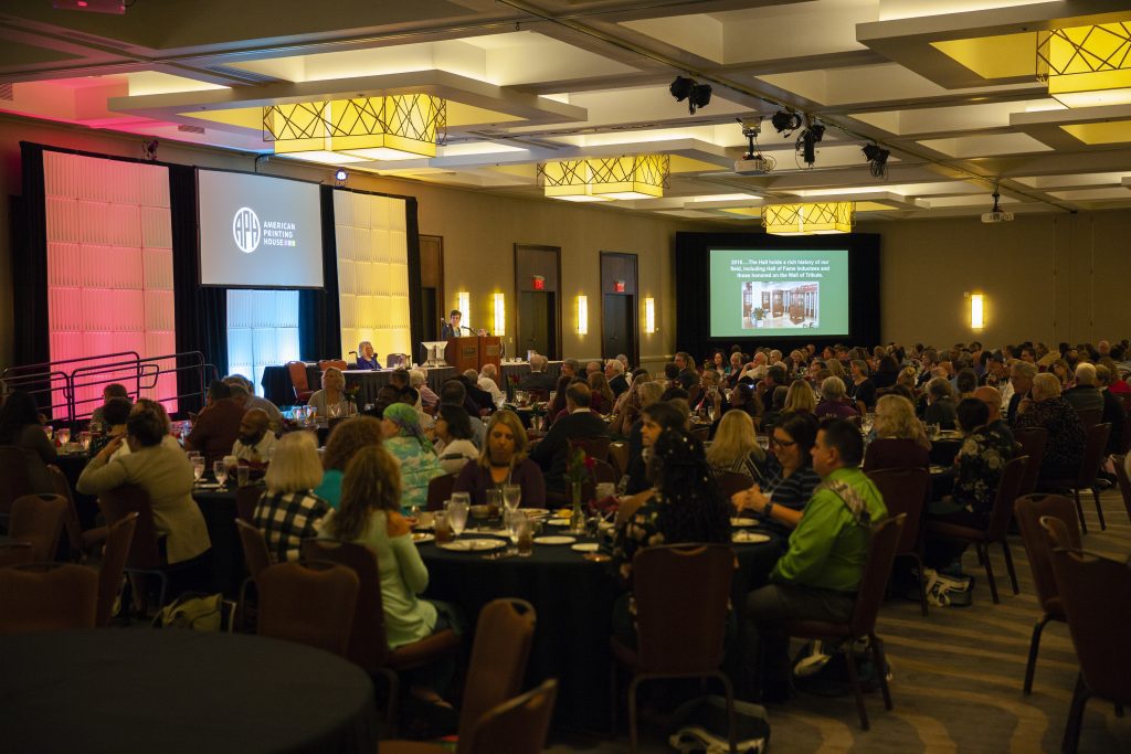 a ballroom full of people sitting at round tables. the wall behind them is lit in APH branding colors of pomegranate, teal, and gold. the aph logo is shown on a screen