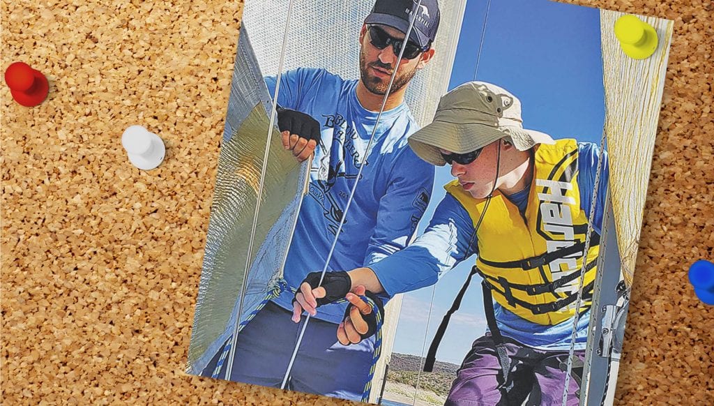 photo pinned to a cork board. photo shows a man in a hat and sunglasses hellping a boy in a lifejacket on a sail boat.