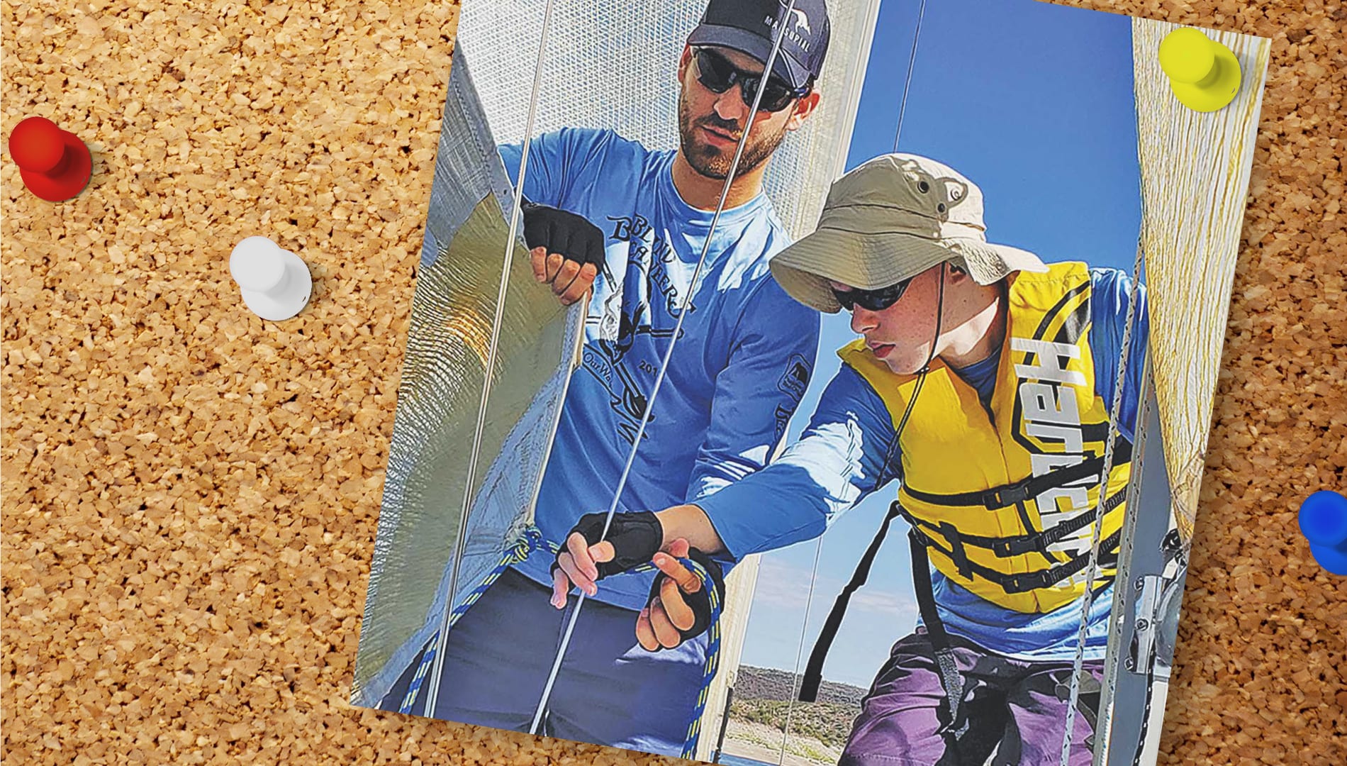 photo pinned to a cork board. photo shows a man in a hat and sunglasses hellping a boy in a lifejacket on a sail boat.