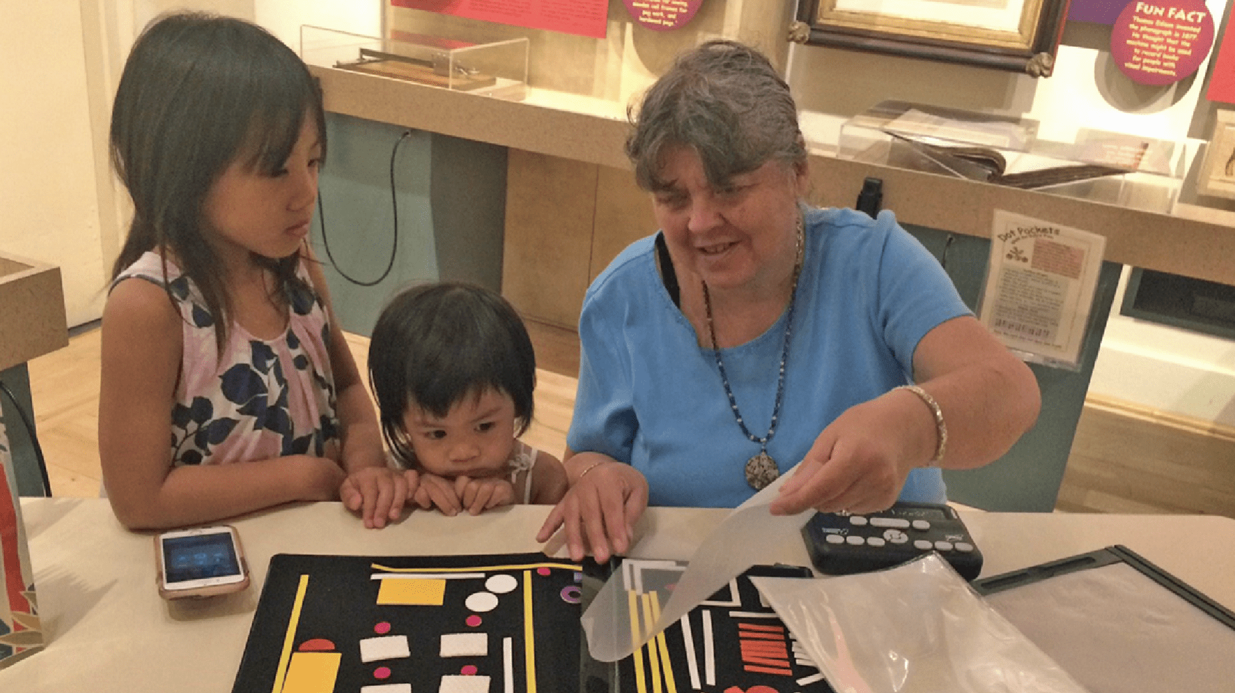 Two children stand next to a woman who is showing them “Picture-Maker,” or the Wheatley Tactile Diagramming Kit