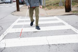 Photo of the legs and feet of a person with a white cane crossing at a cross walk.