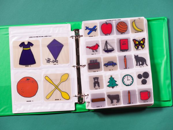 Two pocket pages contained in a green binder with graphic picture cards inserted. On the left side are four large colored picture cards (a blue dress with a yellow sash, a purple kite, an orange, and two yellow spoons crossed), and on the right are twenty small colored picture cards including a blue airplane, a red cardinal, and several other images.