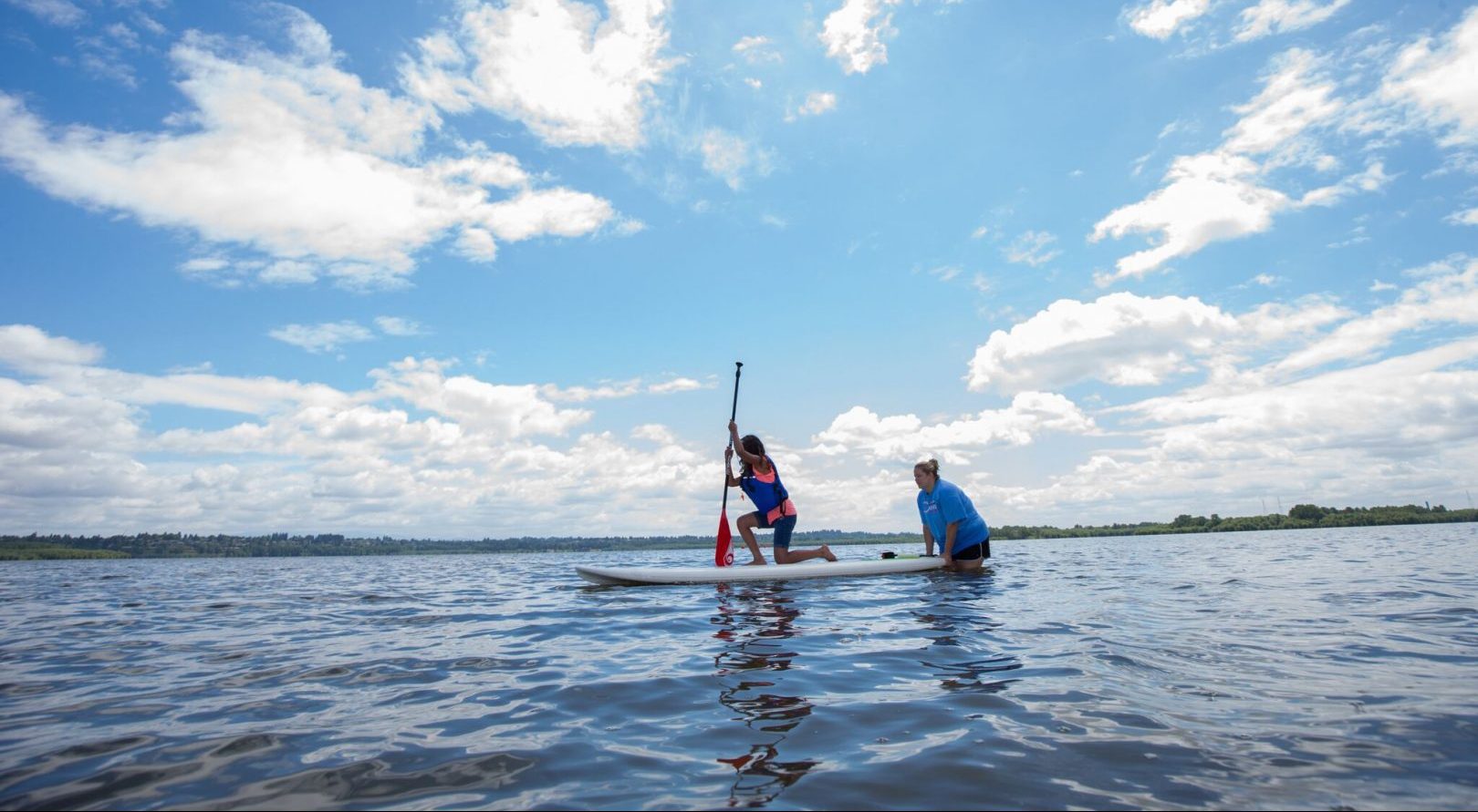 stacey standing in a body of water helping a student on a paddle board