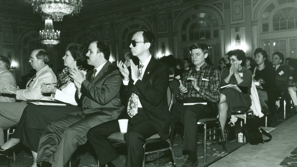 A crowded auditorium. Eight people in the foreground are visible, while the others blur into the background. They are applauding.