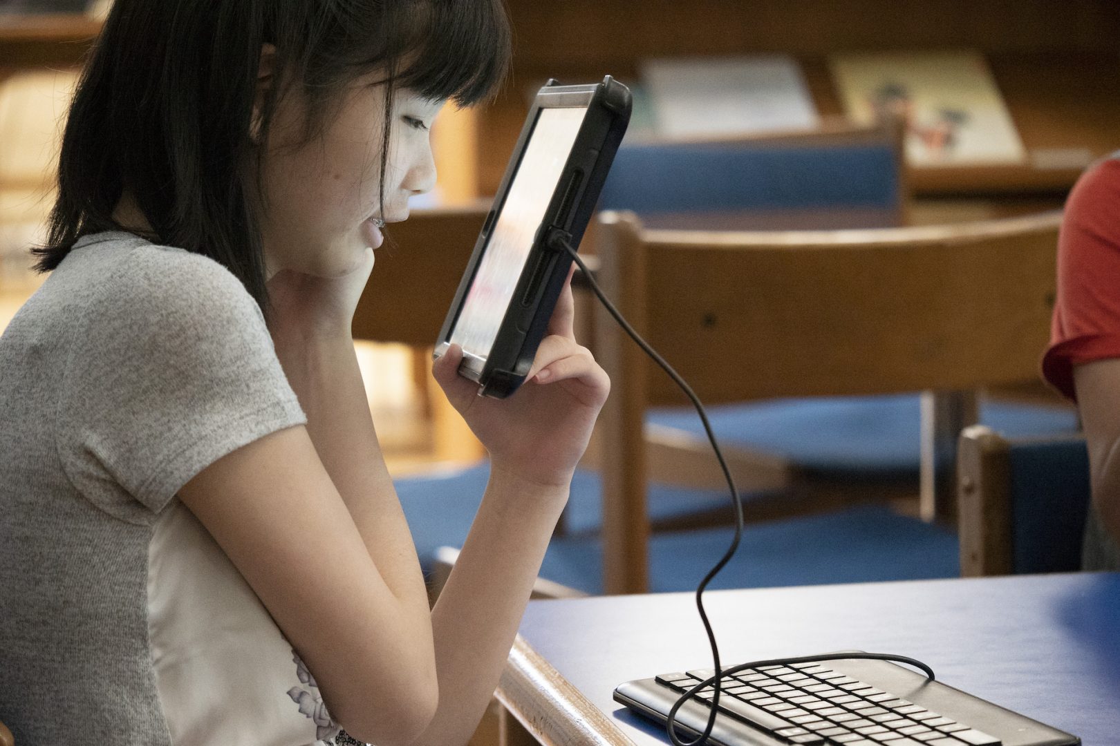 A student sitting at a table smiling while looking up close at a tablet that is connected to a qwerty keyboard