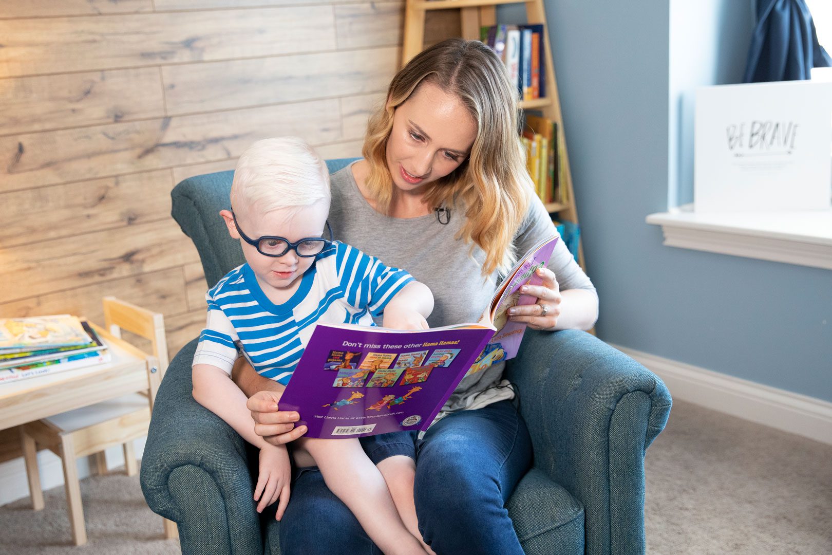 A mother and child sitting in an armchair together wile reading a print/braille book. The child is wearing glasses.
