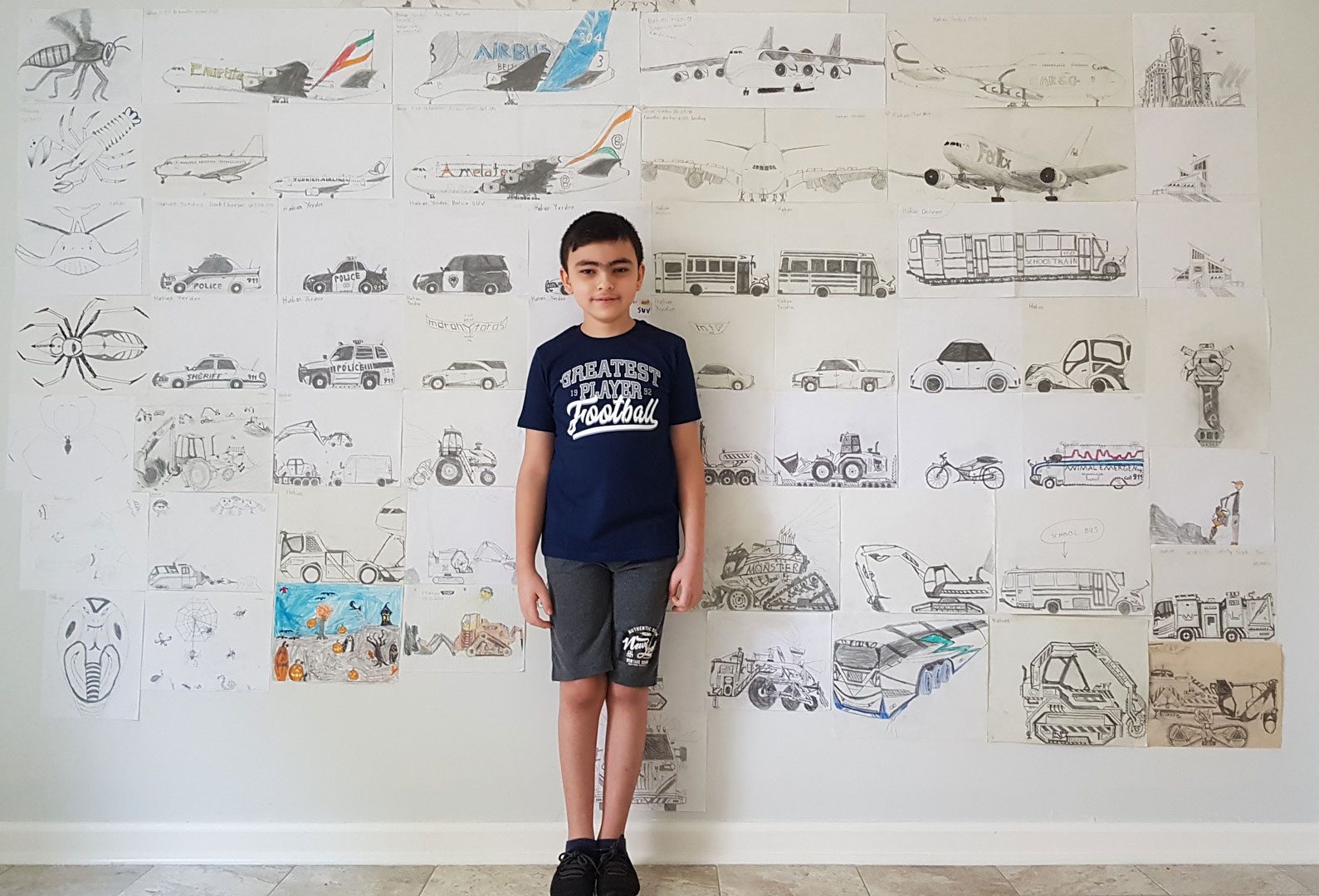 A young Insights Art award winner standing in front of a wall covered in his illustrations of cars, buses, construction vehicles, and insects.