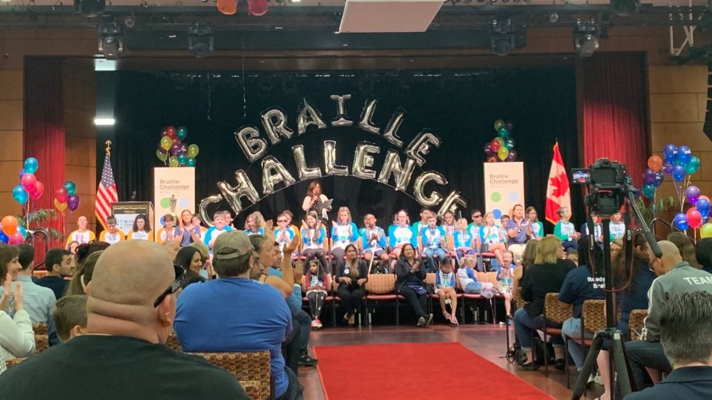 Over 30 students sitting on a stage under large silver balloons spelling out “braille challenge” while the audience watches and claps.