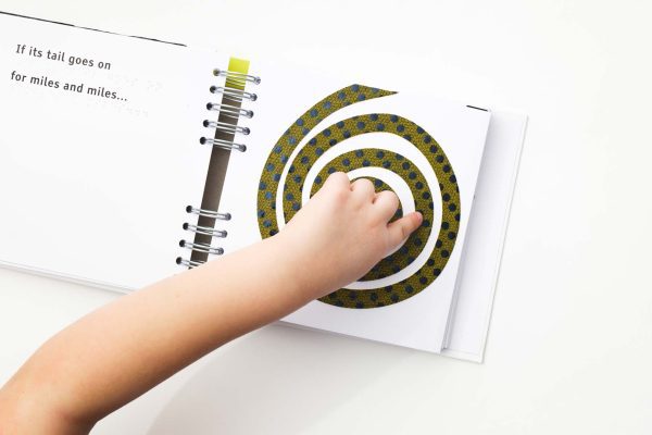 Book opened to show print and braille text “If its tail goes on for miles and miles…” on the left-side page; on the right-side page a child’s finger follows along the monsters long, rolled up tail that is yellow with black textured spots all over it.