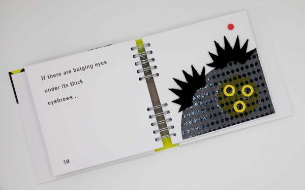 Book opened up to show print and braille text somewhat out from frame. The words that can be read are “…are bulging eyes … its thick…” on left-side page; on the right-side page a child’s hand touches a large black monster covered in a small black spotted texture with thick, spiky, black eyebrows made of felt, and three yellow plastic eyeballs.