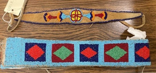 A beaded headband and belt laid out on a table. The headband is a narrow strand of tanned leather with a delicate beaded design in blue yellow, red, gold, and yellow. Dark blue beads run along the boarder. The belt is thicker and completely beaded in light blue beads with squares in colors of blue and maroon. Within those squares are diamond shapes in red and green. 