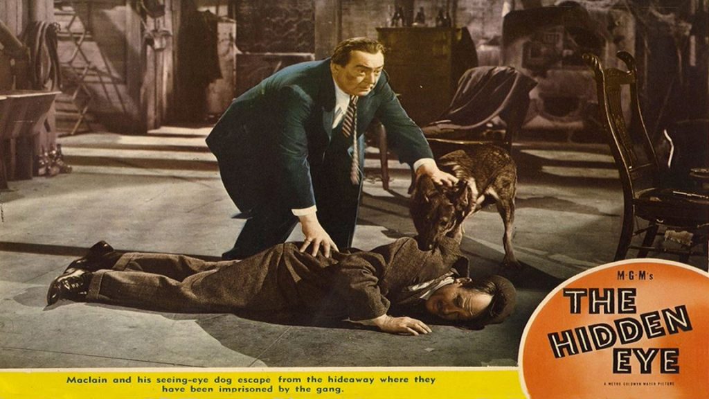 Still image from a movie. A man bends over a body lying on the floor. One of his hands is on the man’s jacket, and the other is reaching toward him. A guide dog sniffs curiously at the body. A banner across the bottoms says 