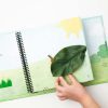 A child’s hand interacting with the fuzzy caterpillar who is underneath a large, green tactile leaf attached to the page.
