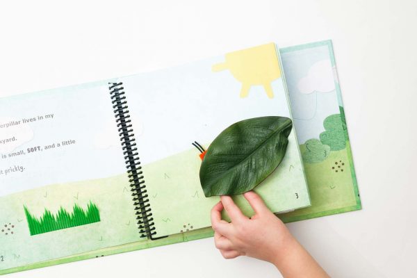 A child’s hand interacting with the fuzzy caterpillar who is underneath a large, green tactile leaf attached to the page.