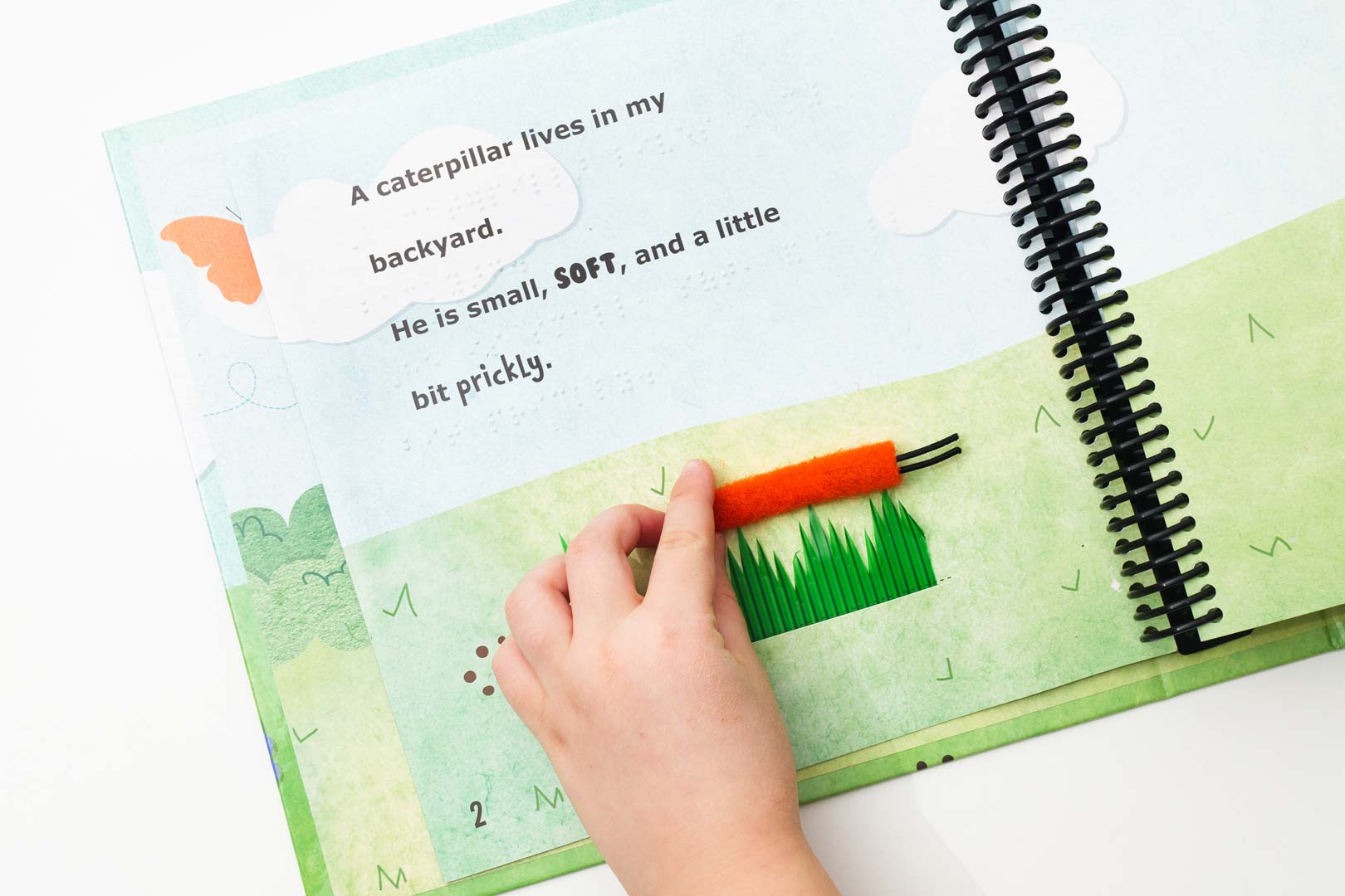 A child uses their hand to move the orange caterpillar across a patch of tall, green grass with the text and braille on the page reading, “A caterpillar in my backyard. He is small, SOFT, and a little bit prickly.”