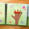 The orange caterpillar made of fuzzy fabric with antennae sits across a foam cut out of child’s hand against a green field.