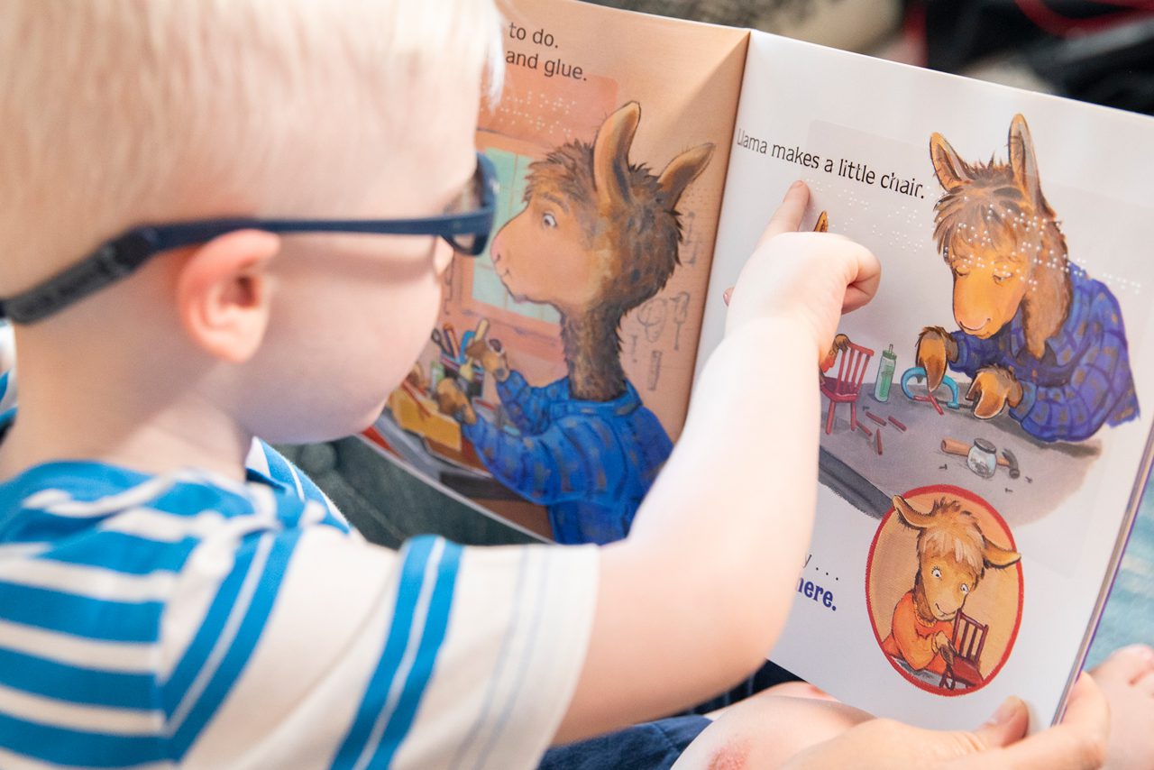 Over the shoulder view of a young child with glasses reading a print/braille book.