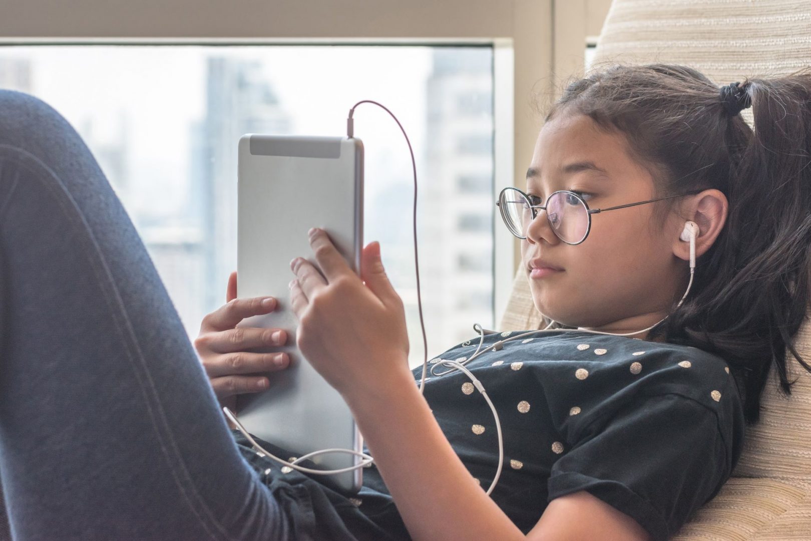 A girl looking at a tablet and wearing earphones while reclining next to a window.