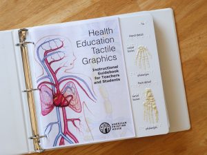 A binder open on a table top. The front page of the Health Education Tactile Graphics sits on top of a tactile graphic of the bones in the hand and foot with text and braille labels.