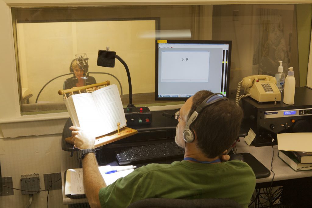 A view into a recording studio. A man in headphones sits in the foreground referencing a print book while the computer next to him counts minutes of recorded material. In the distance, a woman wearing headphones is speaking into a microphone.