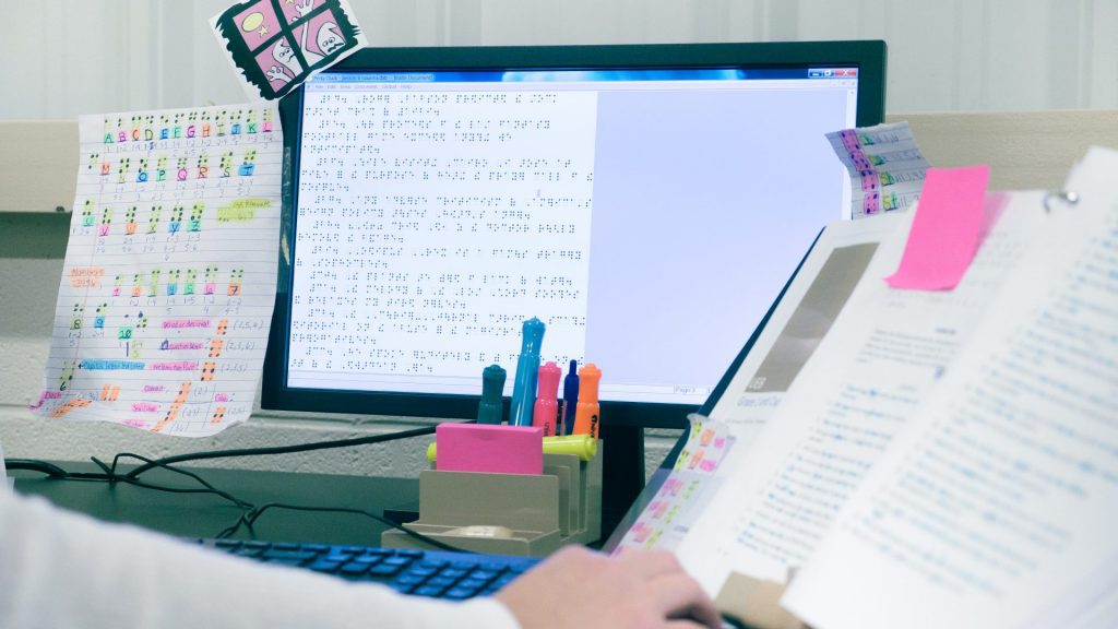 A work station with a computer displaying braille, a desk organizer holding highlighters and Post-It notes, a paper taped to the monitor with color coded notes on the braille alphabet, and a little printed illustrations of funny ghosts. A hand is reaching out to turn the page of a marked up text in a binder.