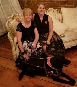 Connie dressed up in a flowery skirt sits with a friend on a beautiful vintage settee. Lexie the black guide dog is wearing a bandana and sits at Connie's feet.