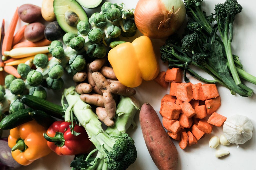 Fresh produce spread across a counter. Includes Brussels sprouts, bell peppers, broccoli, sweet potatoes, and avocados.