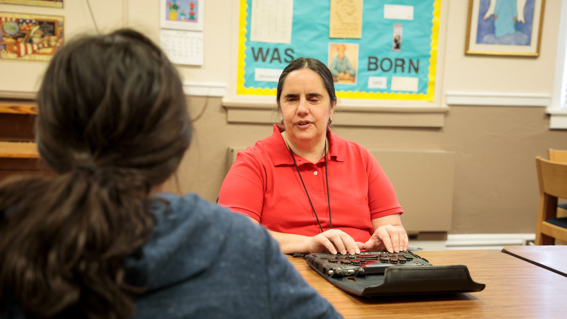 A woman in a red shirt sitting at a table using a braille display. Across from her another person can be seen from the back.