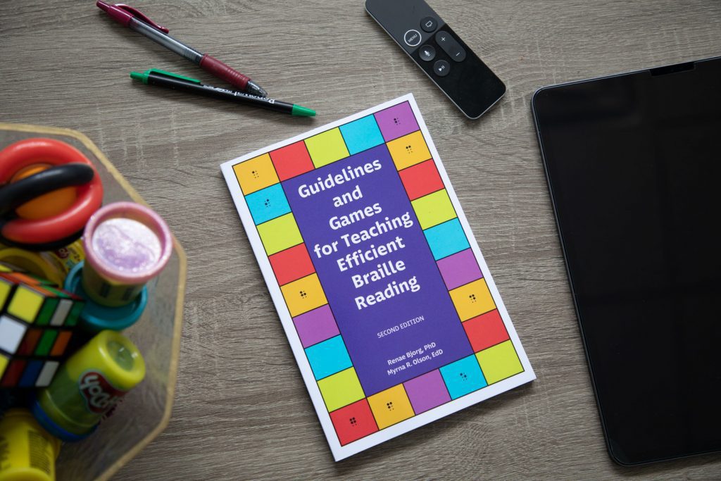 A copy of Guidelines and Games is resting on a dark gray table. To the left of the book is a container holding a Rubik’s Cube and Play-Doh, while two pens and an Apple TV remote are placed above the book, and an iPad is placed on the right side of the book.