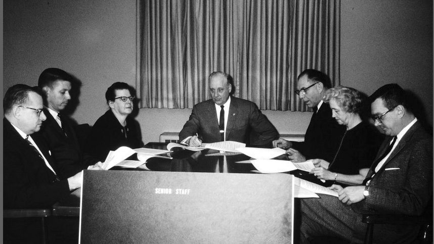 Seven people, five men and two women, are sitting around a table. The men are wearing suits and ties; the women are wearing dark colored dresses. A label reads “Senior Staff.”