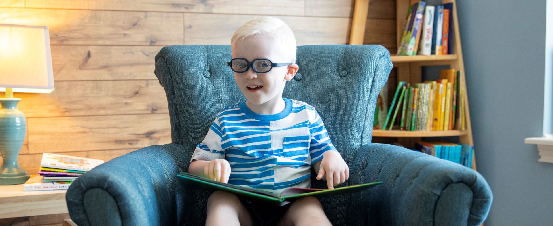 A child wearing glasses sitting in a blue armchair while reading a braille storybook.