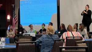 A panel of 8 people sit at the front of a room. A sign language interpreter stands behind them.