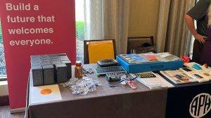 A tabe full of APH products. A pink banner in the background reads "Build a future that welcomes everyone." An APH logo is shown on a table runner.