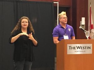 Jeff in a purple APH shirt speaks from behind a podium. An ASL interpreter stands next to him. 