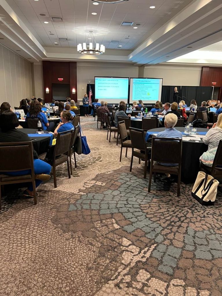 A ballroom filled with round tables. People are seated and looking forward at a presentation on a set of large screens.