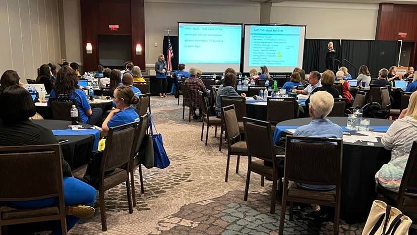 A ballroom filled with round tables. People are seated and looking forward at a presentation on a set of large screens.