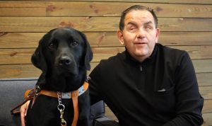 A photo of Italy, a big black lab, sitting next to Richard on a couch. 