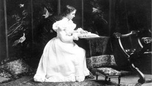 Studio photo of Helen Keller as a teenager. She wears a long white dress with a full skirt and puffed sleeves. White lace decorates the high neck, the sleeves, and the edge of the skirt.