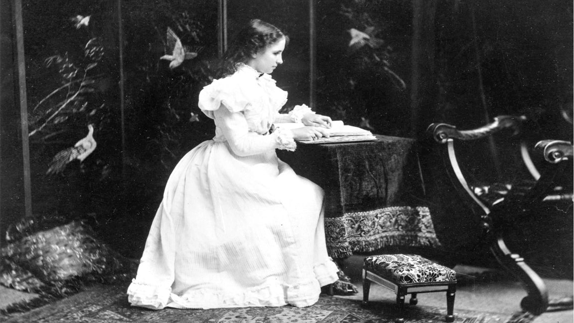 Studio photo of Helen Keller as a teenager. She wears a long white dress with a full skirt and puffed sleeves. White lace decorates the high neck, the sleeves, and the edge of the skirt.