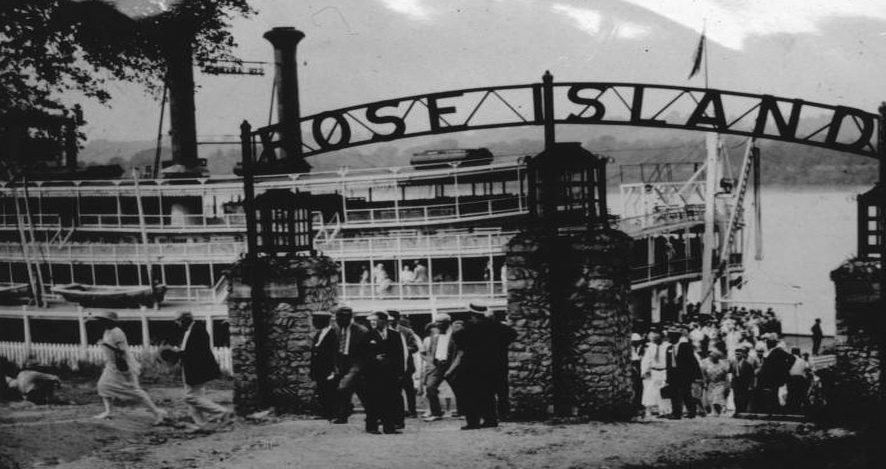 People exiting a large steamboat and passing under a large metal sign reading “Rose Island,” which is held up by three large stone pillars.