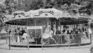 Fenced-in carousel tightly packed with people dressed formally and carousel horses. The gap in the fencing in front of the carousel has a sign reading “entrance” above it. There are many trees behind the carousel.